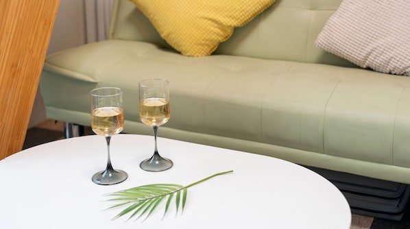 Dos copas de vino blanco sobre una mesa blanca con una decoración de hoja de palma, frente a un sofá verde con un cojín amarillo.