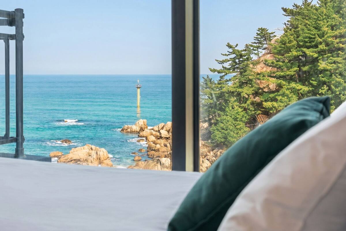 A bedroom with a large window overlooking a rocky coastline and a lighthouse.