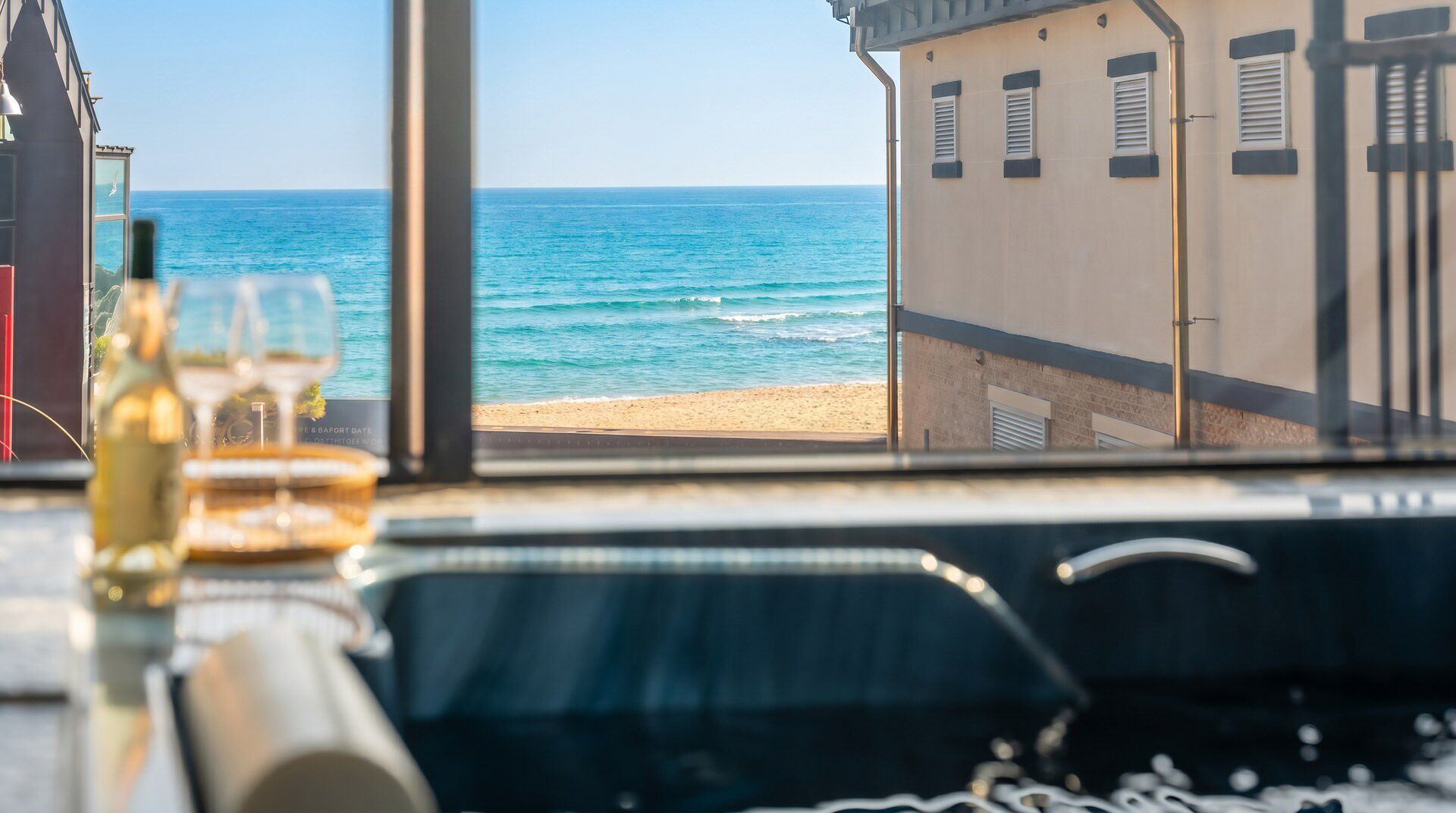 A view from a window overlooking a beach, with a table in the foreground containing a bottle and a glass.