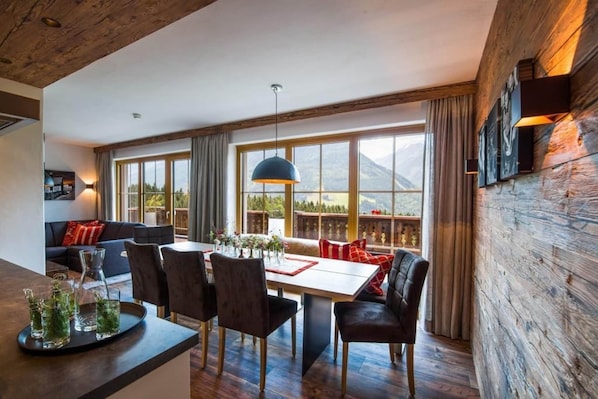 Uma sala de jantar com uma mesa de madeira, cadeiras e vista para montanhas através de grandes janelas.