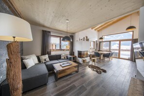 Uma sala de estar moderna com um sofá cinza, mesa de centro de madeira e lareira. Há janelas amplas com vista para as montanhas.