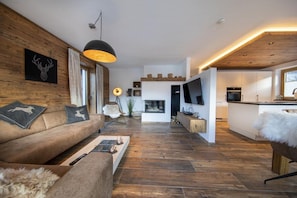 Una sala moderna con piso de madera, un sofá con almohadones de reno, una chimenea y una zona de cocina con un televisor grande.