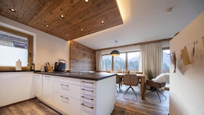 A modern kitchen with wooden ceiling, white cabinets, and a large island. There is a dining area with a table and chairs, and a view of mountains through the window.