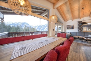 Un tavolo da pranzo in legno con tovaglietta a scacchi, sedie rosse rivestite in tessuto e vista sulle montagne innevate attraverso ampie finestre.