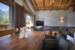 Uma mesa de jantar de madeira com um vaso de flores, um sofá e uma televisão num quarto com teto de madeira e janelas amplas.