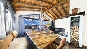 Un comedor espacioso con una gran mesa de madera y sillas de cuero, una zona de estar acogedora con una manta de piel, y vistas a montañas a través de grandes ventanales.