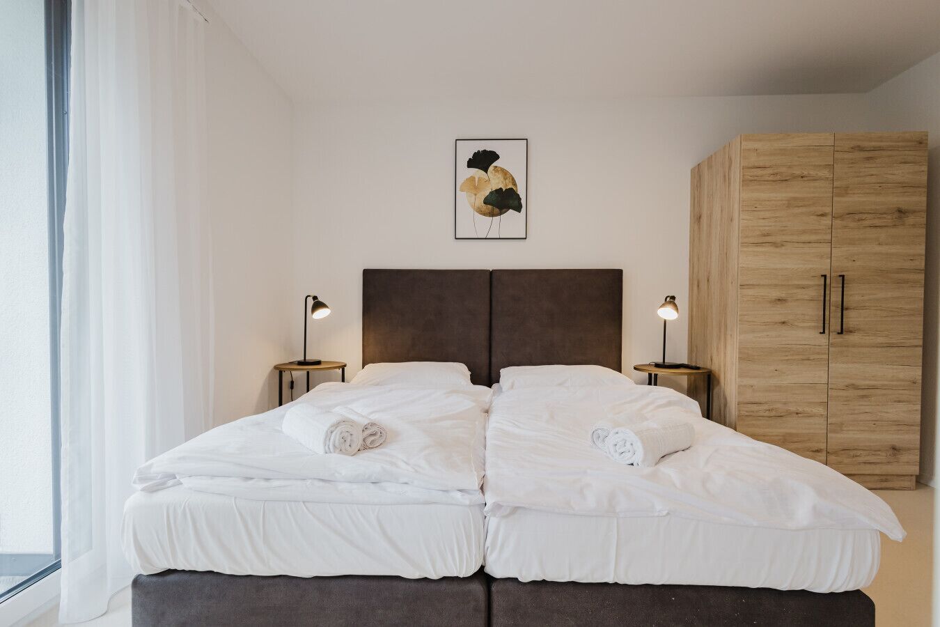 Un lit double avec du linge de lit blanc, deux tables de chevet avec des lampes, une armoire en bois et une œuvre d’art encadrée au mur.