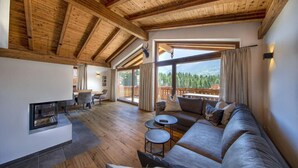 A spacious living room with a wooden ceiling, a fireplace, a large sofa, and a view of a forested mountain. - Chalet with sauna, Neukirchen (Neukirchen)