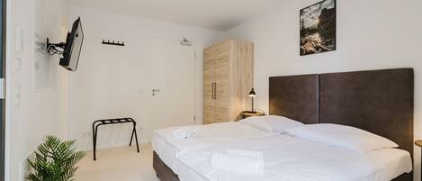 Un dormitorio moderno con una cama grande, un armario de madera, una mesita de noche pequeña, un televisor montado en la pared y una planta en maceta.