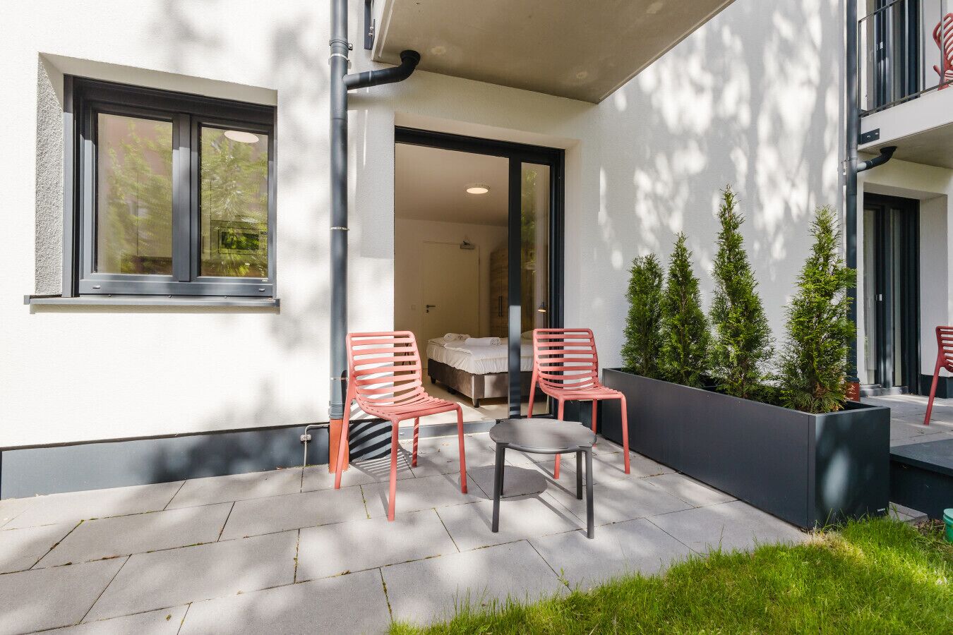 A modern outdoor seating area with two red chairs and a table, a small garden with potted plants, and a building entrance with a glass door.