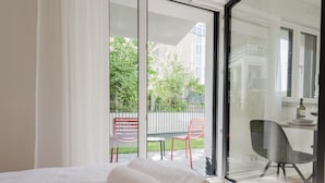 A hotel room with a bed, a desk with a chair, and a view of a balcony with chairs and a fence. - The InnSight - Apartmenthouse (Passau)
