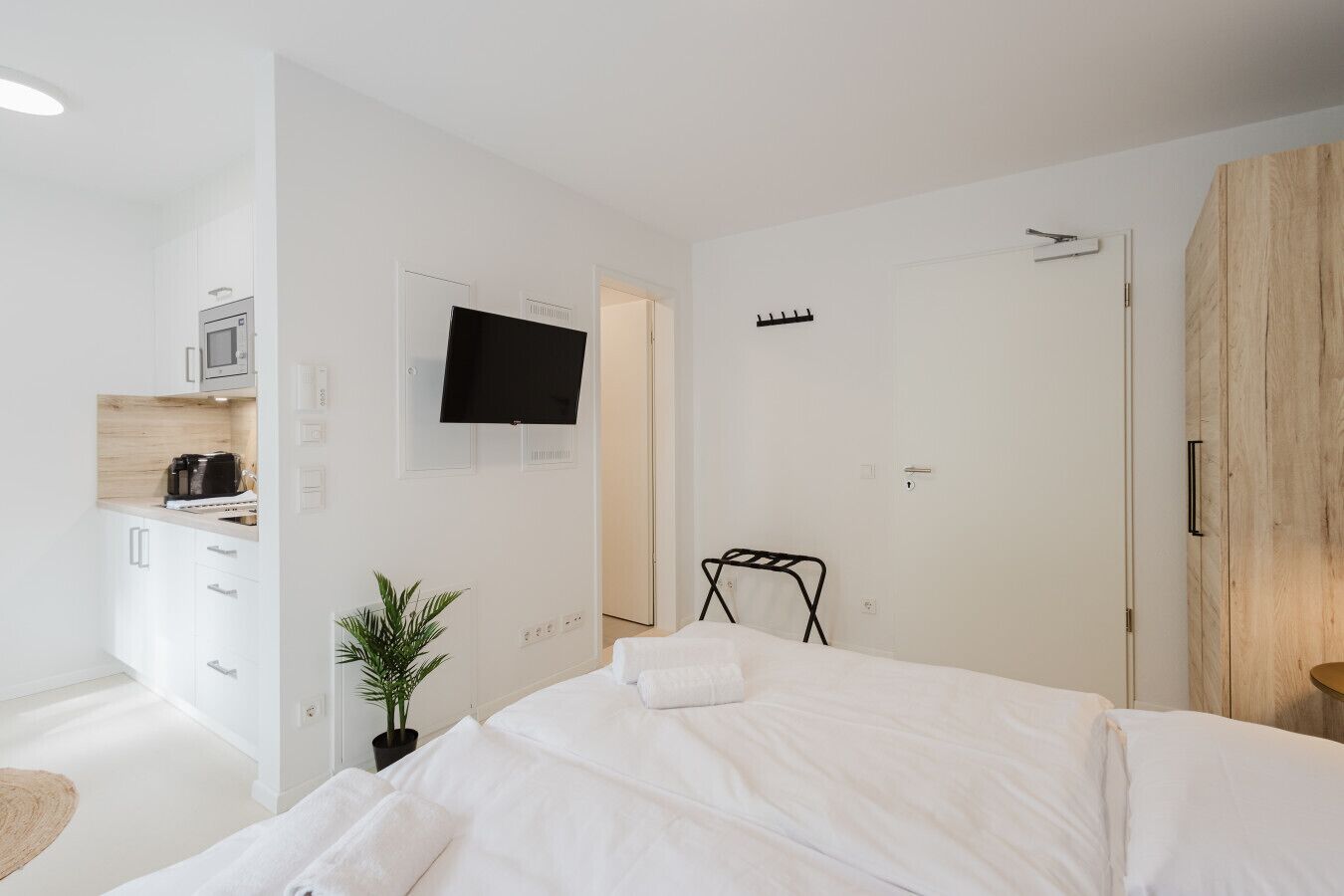 A modern hotel room with a bed, a TV mounted on the wall, a small kitchenette, and a wooden wardrobe.