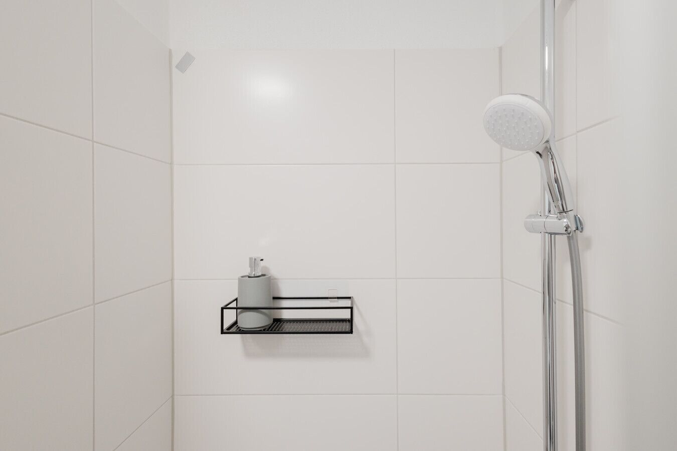 En dusch med ett duschhuvud som kan hållas i handen, en väggmonterad tvåldispenser och en tvålkopp.