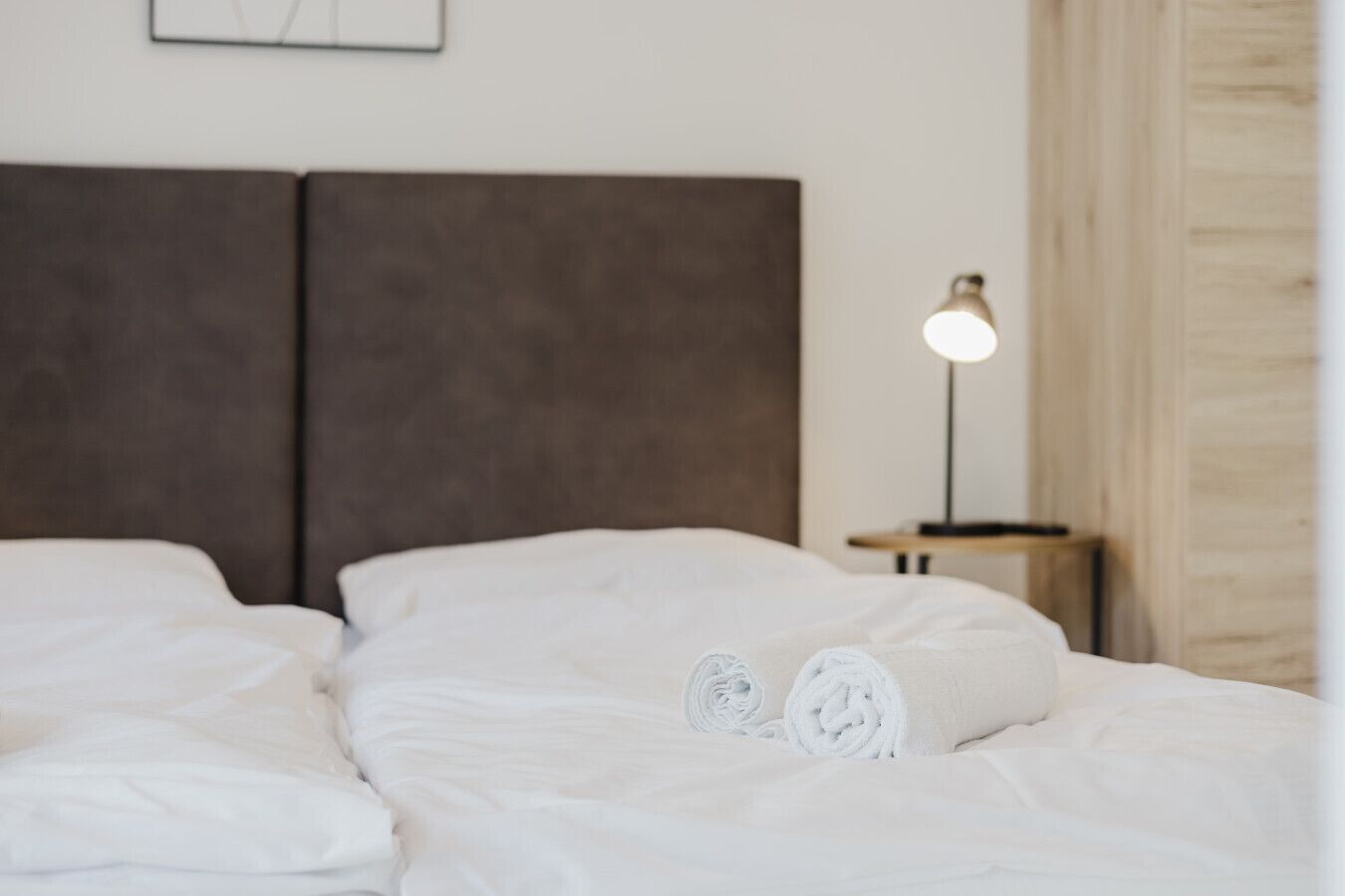 A neatly made bed with white linens and rolled towels, a modern headboard, and a bedside lamp on a wooden nightstand.