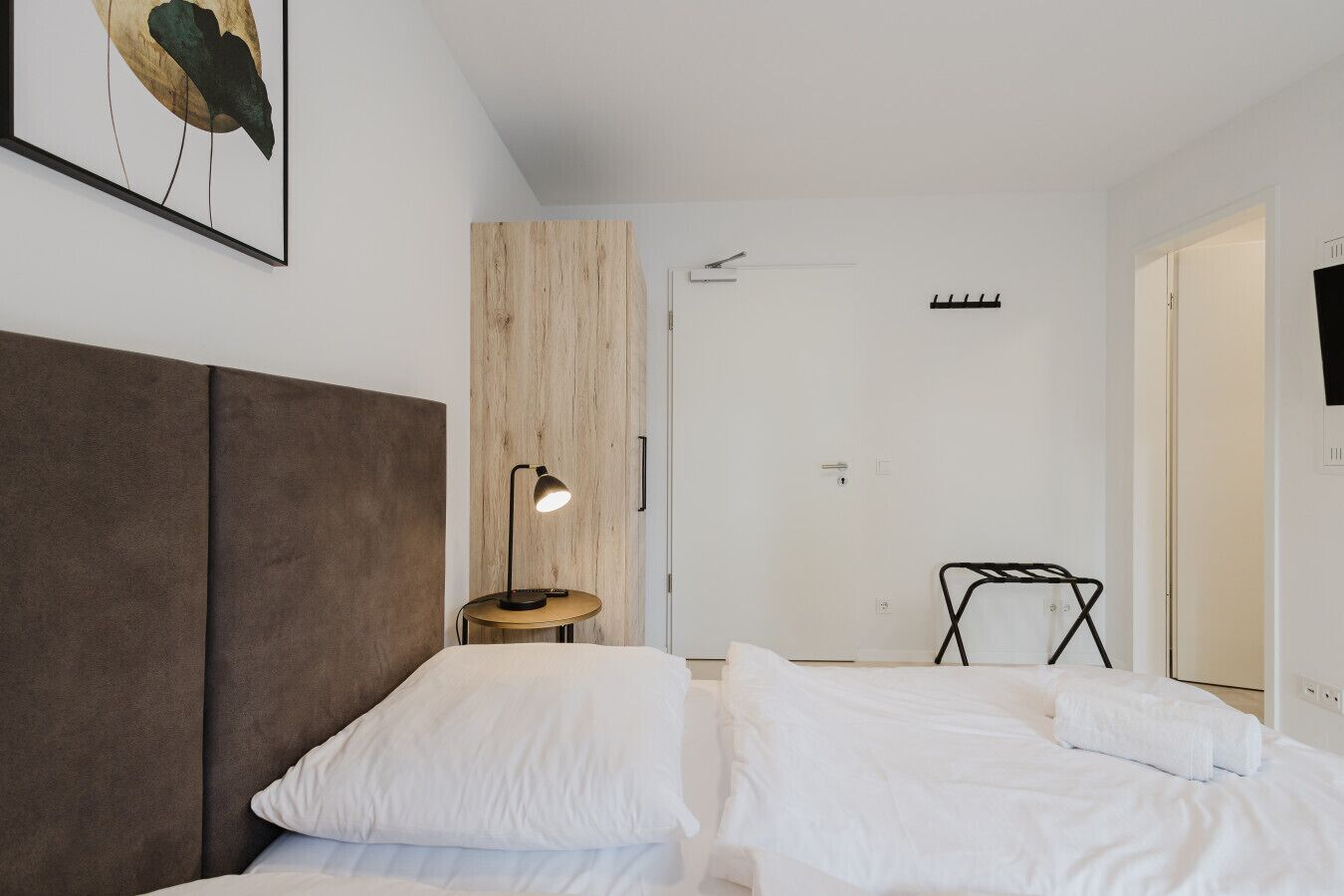 A modern hotel room with a large bed, a bedside table with a lamp, a wooden accent wall, and a door with a coat rack.