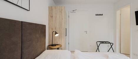 A modern hotel room with a large bed, a bedside table with a lamp, a wooden accent wall, and a door with a coat rack.