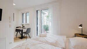 A modern hotel room with a bed, a desk, and a chair. There is a window with curtains, a lamp, and a view of the outside. - The InnSight - Apartment house (Passau)