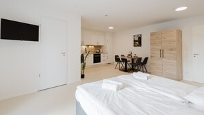 Une chambre moderne avec un grand lit, un coin repas avec une table et des chaises, une cuisine à l’arrière-plan et un téléviseur fixé au mur.