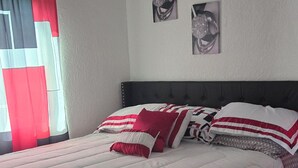 A bedroom with a bed, a window with curtains, an air conditioning unit, and two wall-mounted pictures.