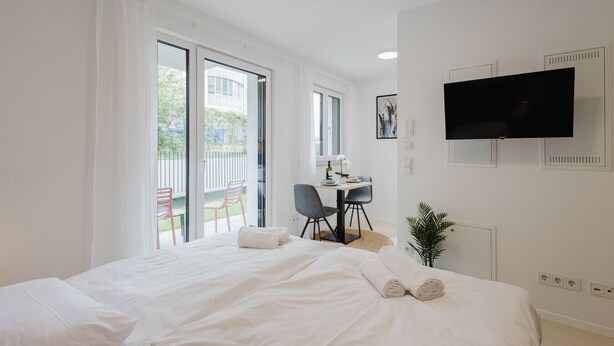 A hotel room with a bed, a desk, a chair, a TV, and a view of the outside. - The InnSight - Apartmenthouse (Passau)