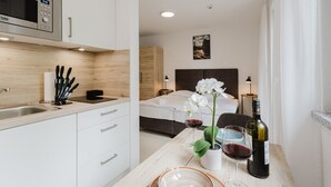 A modern hotel room with a kitchenette, a bed with a headboard, a bedside table with wine glasses and a bottle, and a view of the room. - The InnSight - Apartmenthouse (Passau)