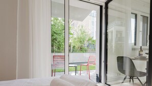 A hotel room with a bed, a desk with a chair, and a view of a balcony with chairs and a fence. - The InnSight - Apartmenthouse (Passau)