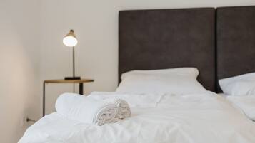 Ein ordentlich gemachtes Bett mit weißen Leinen und einem gerollten Handtuch, ein Nachttisch mit Lampe sowie ein dunkles Kopfteil.
