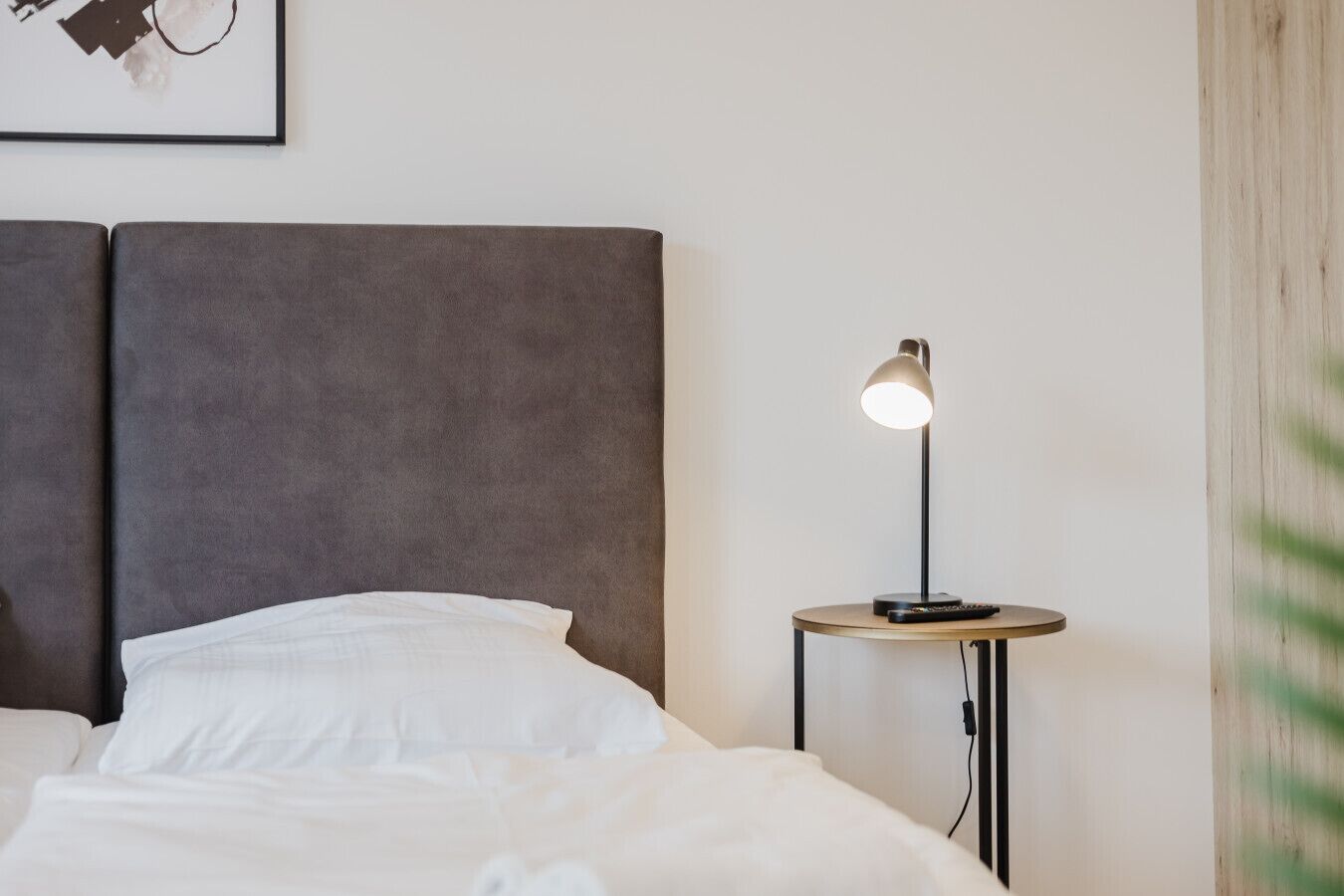 Una cama bien hecha con sábanas blancas, un cabecero tapizado en gris y una mesita de noche con una lámpara y un control remoto.