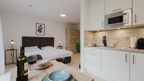 A modern hotel room with a bed, bedside tables, a dining table with wine and dinnerware, a microwave, and a sink. - The InnSight - Apartmenthouse (Passau)