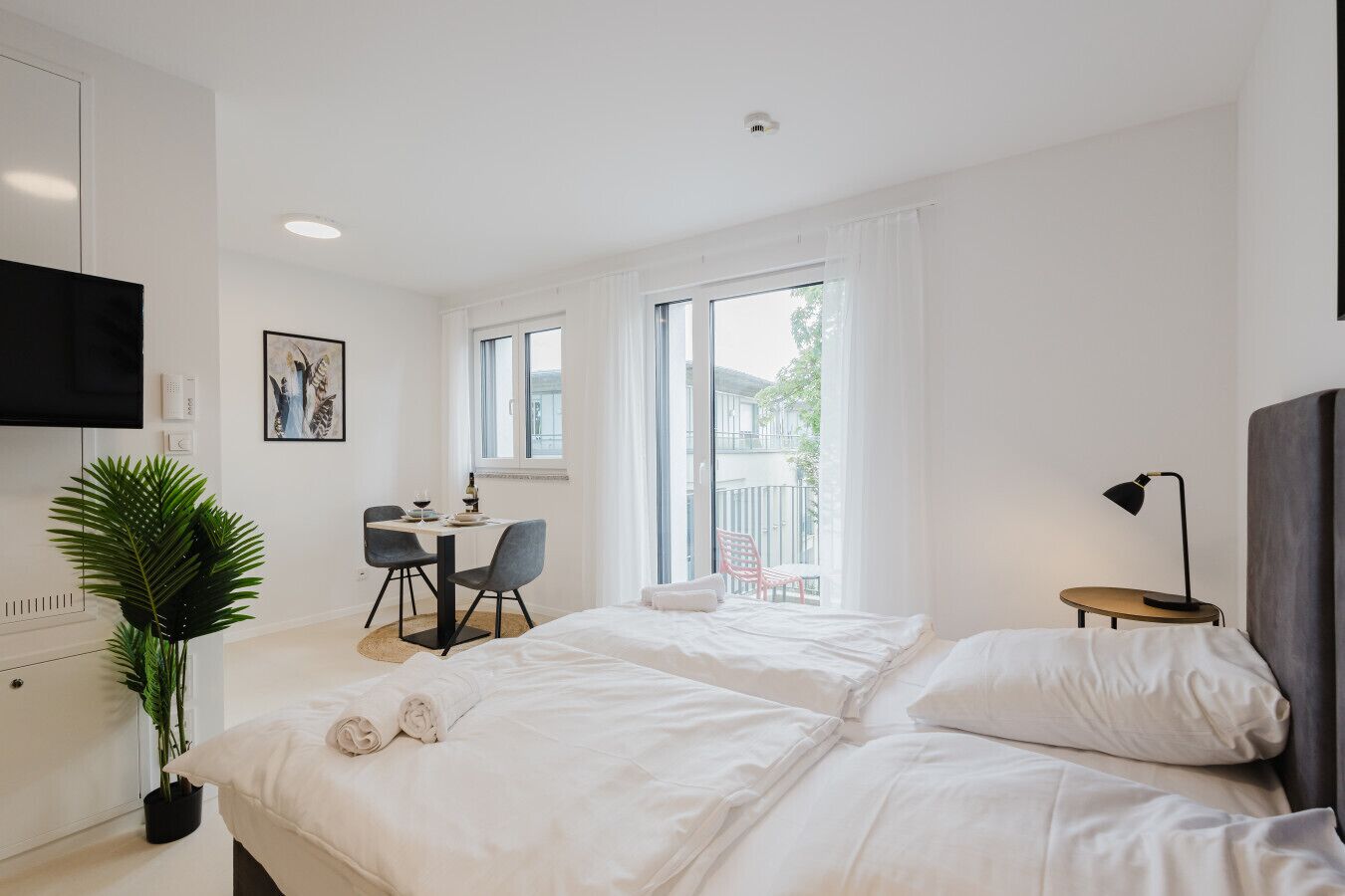 A modern hotel room with a bed, a desk, a chair, a potted plant, and a window with a view of the outside.