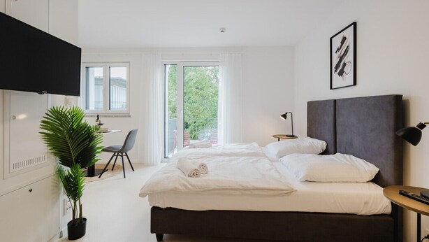 A modern bedroom with a large bed, a desk, and a TV. - The InnSight - Apartmenthouse (Passau)