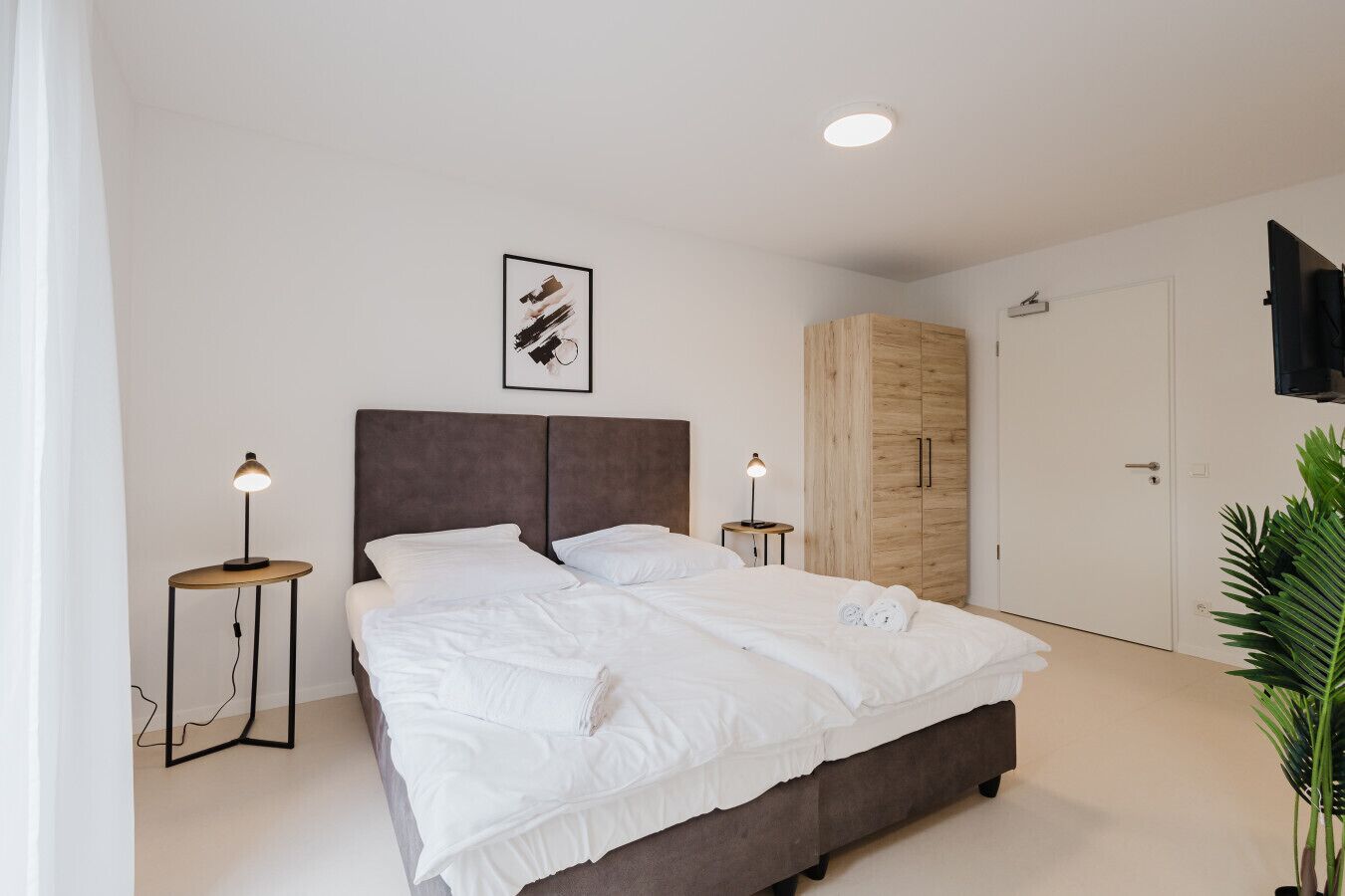 Ein modernes Schlafzimmer mit einem großen Bett, zwei Nachttischen mit Lampen, einem Holzschrank und einer Deckenleuchte.