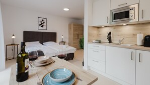 A modern hotel room with a bed, bedside tables, a dining area with a table set for two, a microwave, and a sink. - The InnSight - Apartmenthouse (Passau)