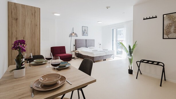 A modern, well-lit room with a dining area, a red chair, a bed, and a view of the city.
