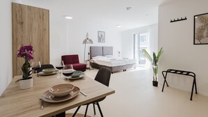 A modern, well-lit room with a dining table set for two, a red sofa, a bed with a grey headboard, and a large window providing natural light. - The InnSight - Apartmenthouse (Passau)