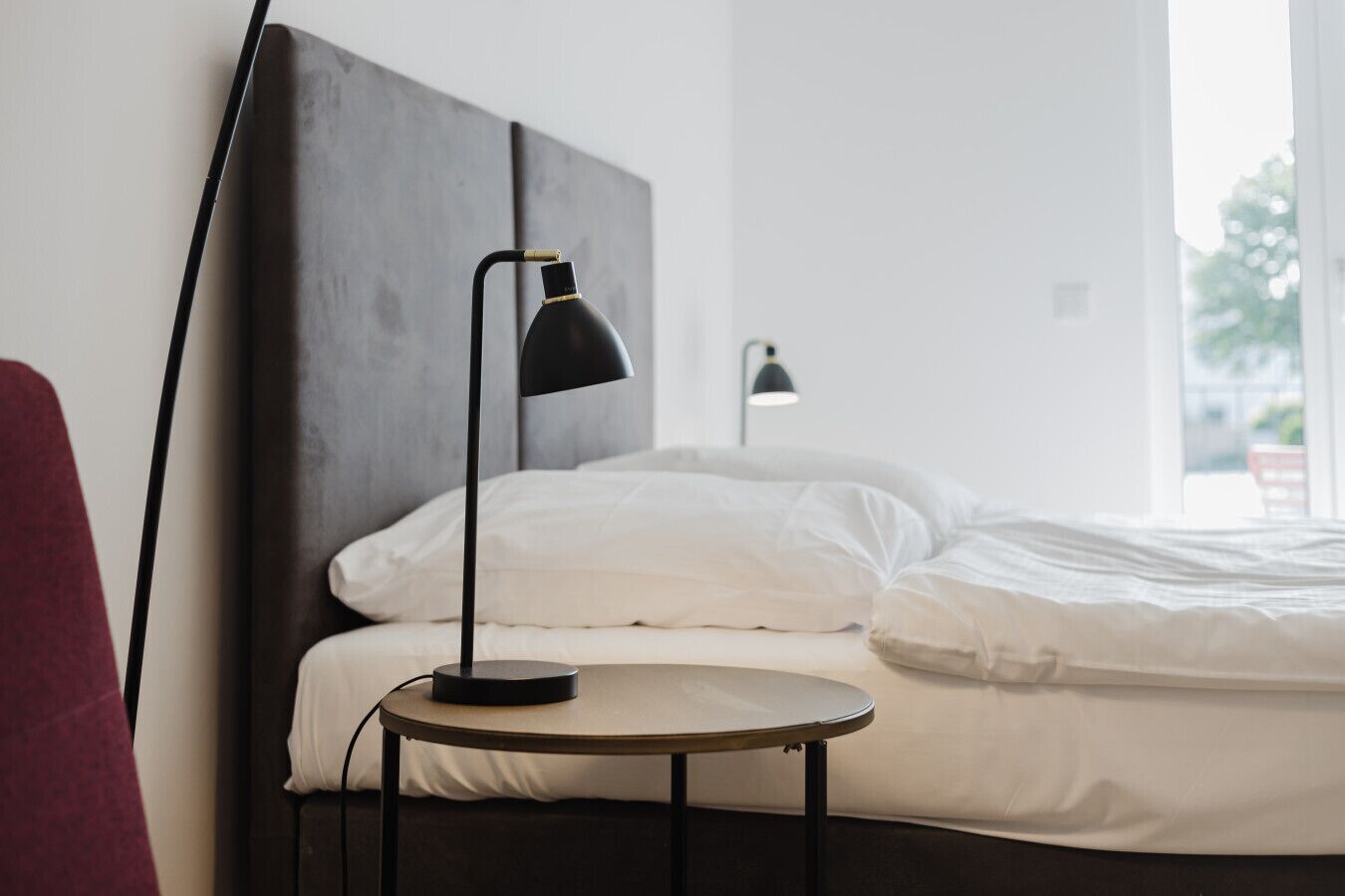 Una cama bien tendida con sábanas blancas, una mesita de madera redonda con una lámpara negra y un cabecero gris.