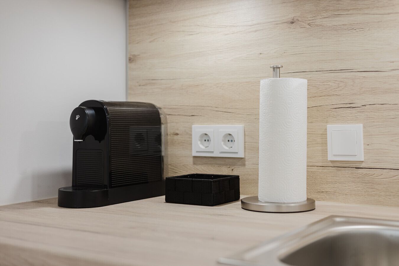 A coffee machine, a paper towel dispenser, and a black trash bin on a wooden countertop with a sink.