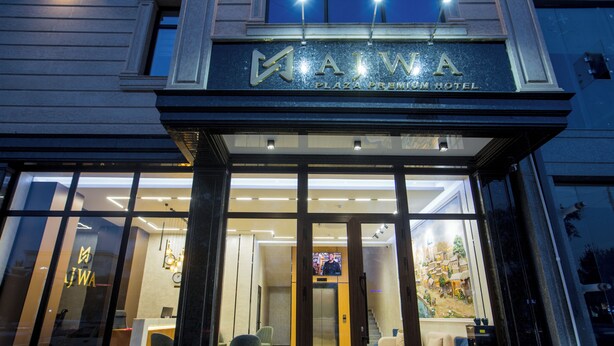 El exterior del Hotel AJWA Plaza Premium, con una fachada moderna, amplias ventanas de vidrio y un mostrador de recepción visible en el interior.
