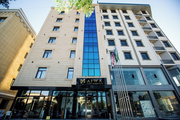 A multi-story hotel with a prominent blue glass facade, a visible entrance with a sign, and a flag outside. - Ajwa Plaza Premium Hotel (Bukhara)