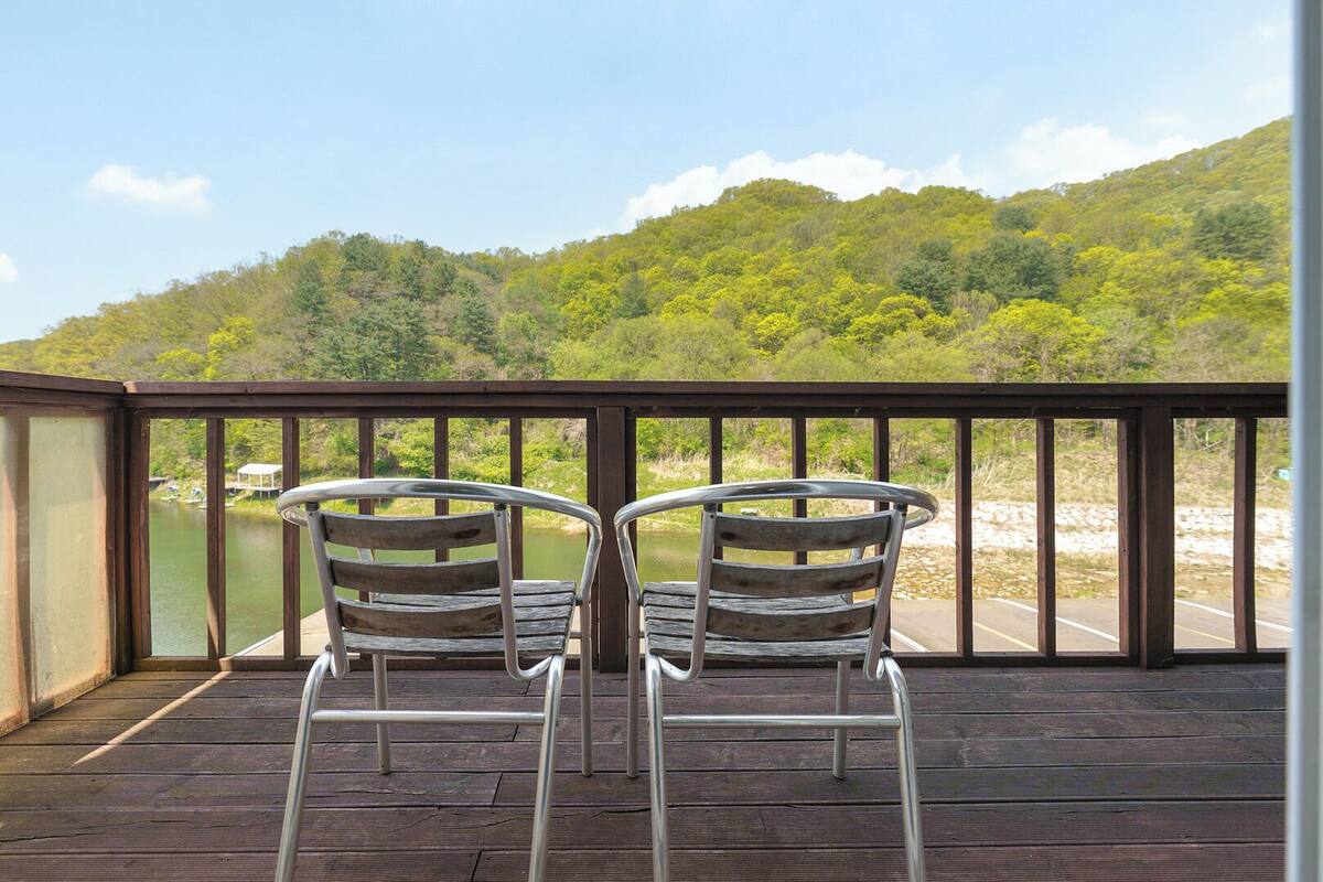 Una terraza de madera con dos sillas de metal que dan a un lago y colinas boscosas.