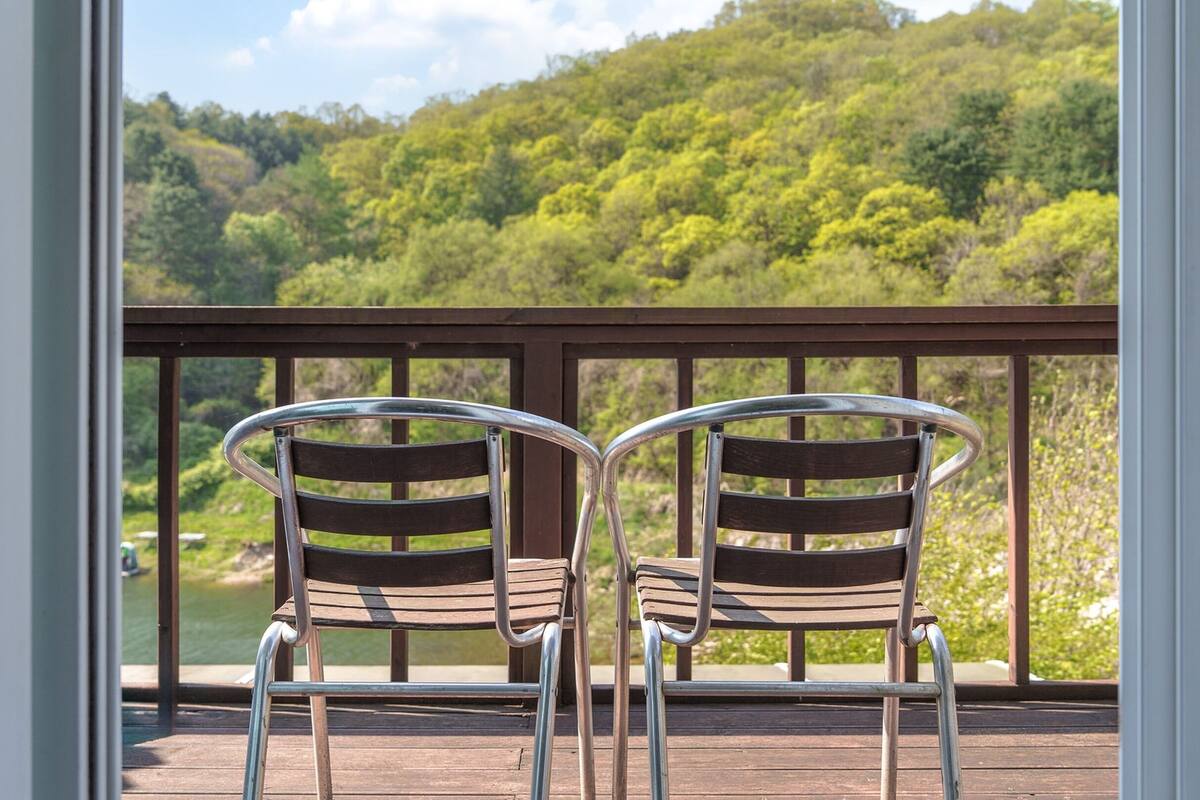 Dos sillas de metal con listones de madera en una terraza de madera con barandilla de metal, con vistas a una ladera boscosa.