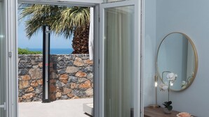 A bedroom with a bed, a wooden dresser, a mirror, and a view of the ocean through an open door. - La Mer de Crete Suite private pool & sea view (Agia Pelagia)