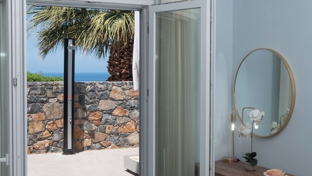 A bedroom with a bed, a wooden dresser, a mirror, and a view of the ocean through an open door. - La Mer de Crete Suite private pool & sea view (Agia Pelagia)