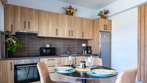 A modern kitchen with wooden cabinets, a built-in oven, a microwave, and a refrigerator. There is a dining table with chairs and a vase of flowers. - La Mer de Crete Suite private pool & sea view (Agia Pelagia)