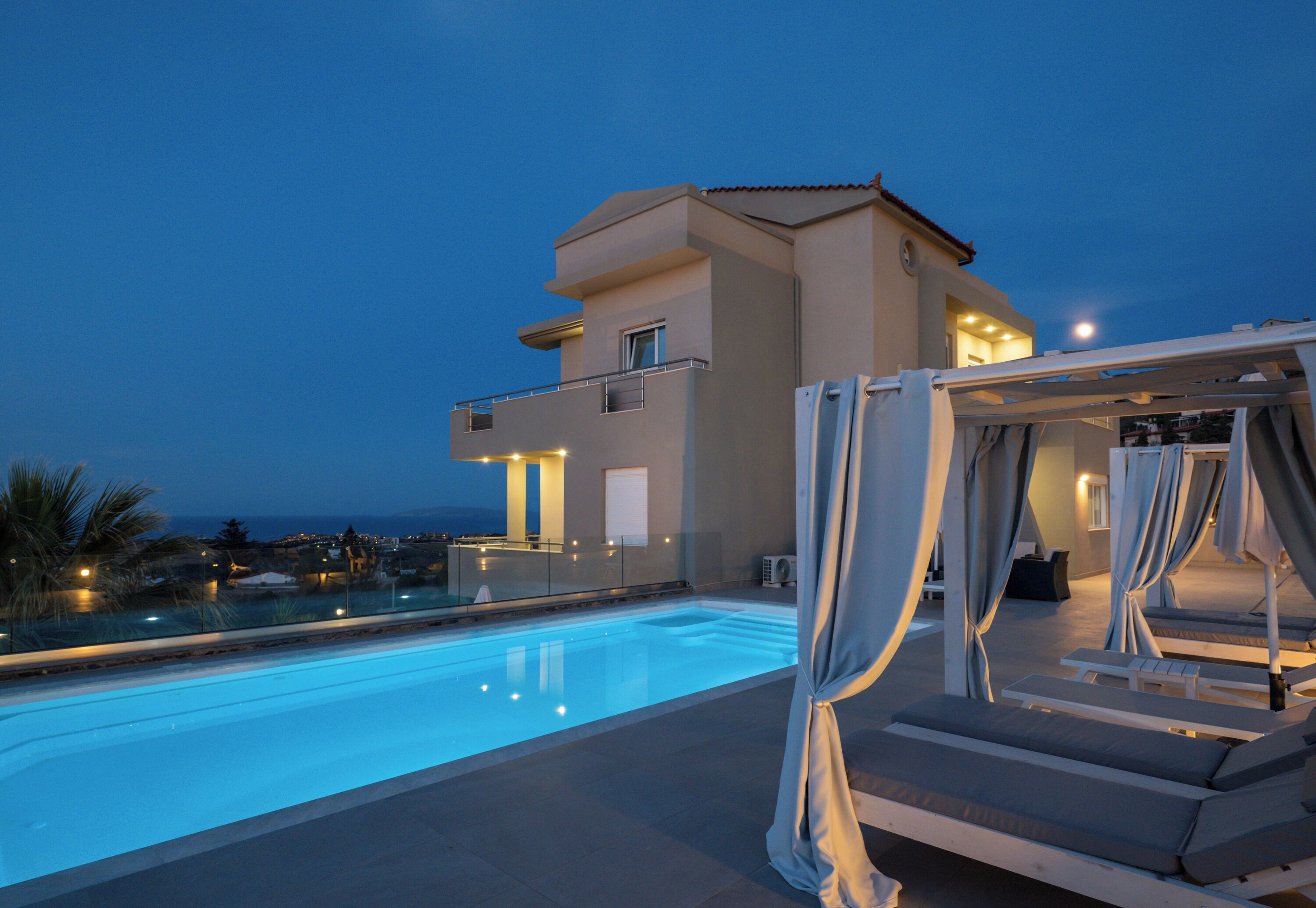 A rooftop pool area with a clear view of the sea, a two-story building, and a covered seating area with curtains.