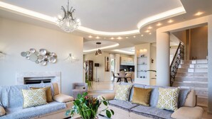 A modern living room with a sofa set, a coffee table, and a chandelier. There is a kitchen in the background with a dining area. - Villa La mer de Crete - private pool & sew view (Agia Pelagia)