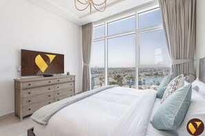 A bedroom with a large bed, a dresser, a TV, and a view of a cityscape through large windows.
