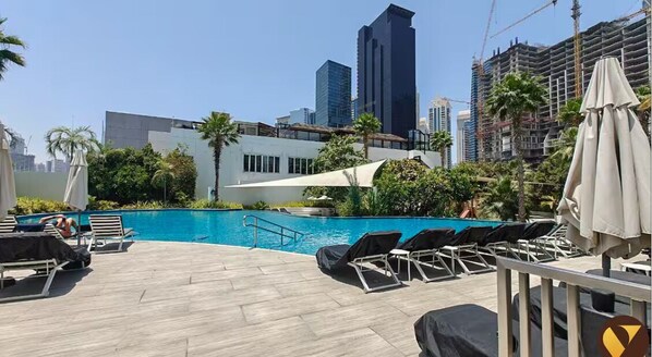 A rooftop pool area with lounge chairs and umbrellas, surrounded by palm trees and modern buildings.