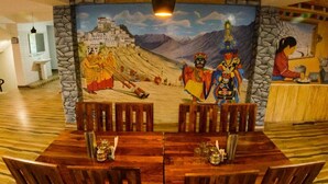 A dining area with a mural depicting a traditional scene, a wooden table with chairs, and a stone wall. - Kapoor Resort Kaza by Mountain Bee (Kaza)
