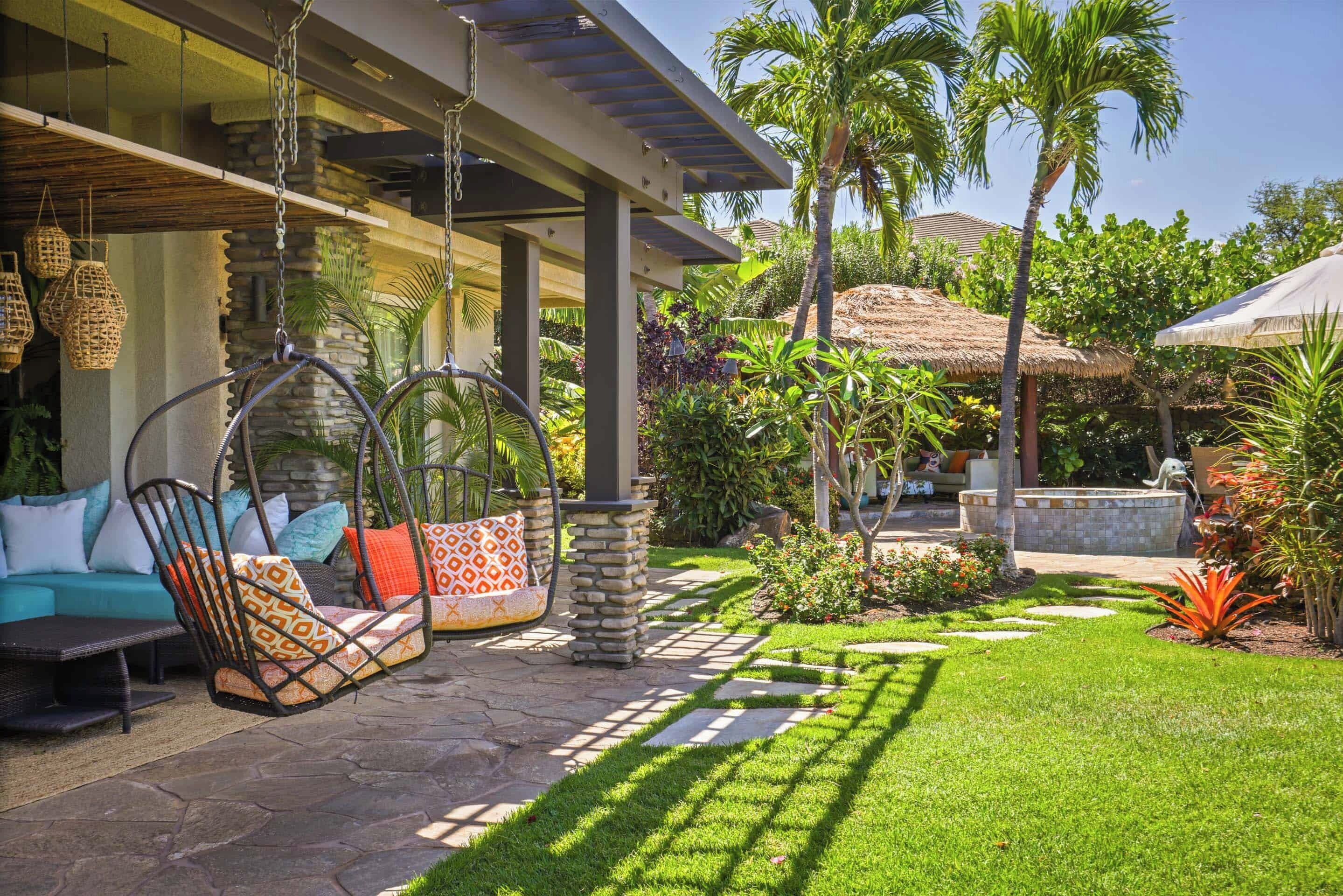 Un patio con sillas colgantes, un sendero de piedra y abundante vegetación, incluyendo palmeras y plantas con flores.
